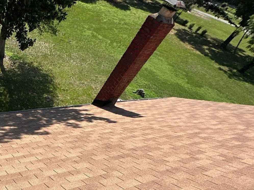 Aging tan three-tab shingles running up to the red brick chimney, lawn and trees beyond