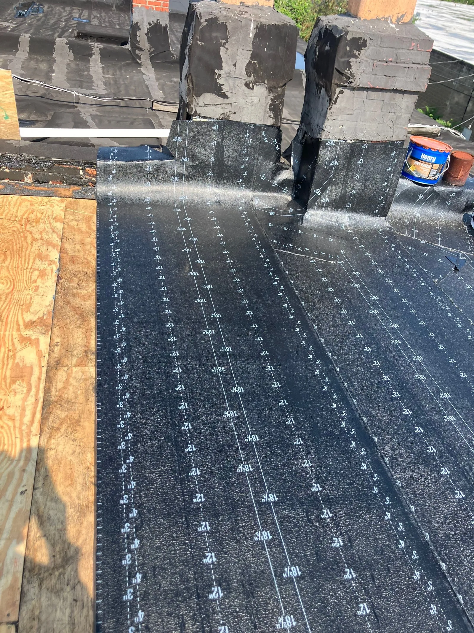 Base sheet rolled out over fresh plywood on a low-slope roof, the old roofing and a chimney with a pipe boot beyond it