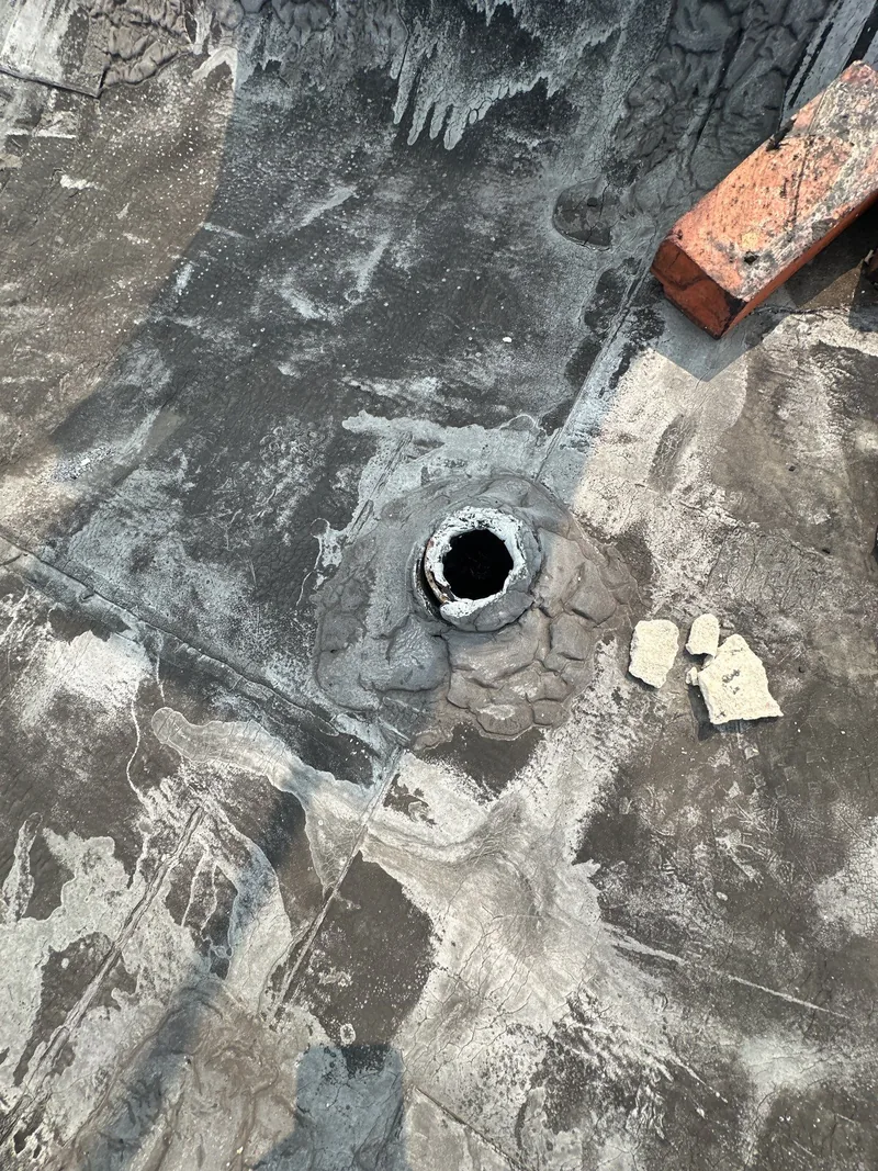 An open pipe penetration in the old coated flat roof surface, loose bricks and a cap lying beside it