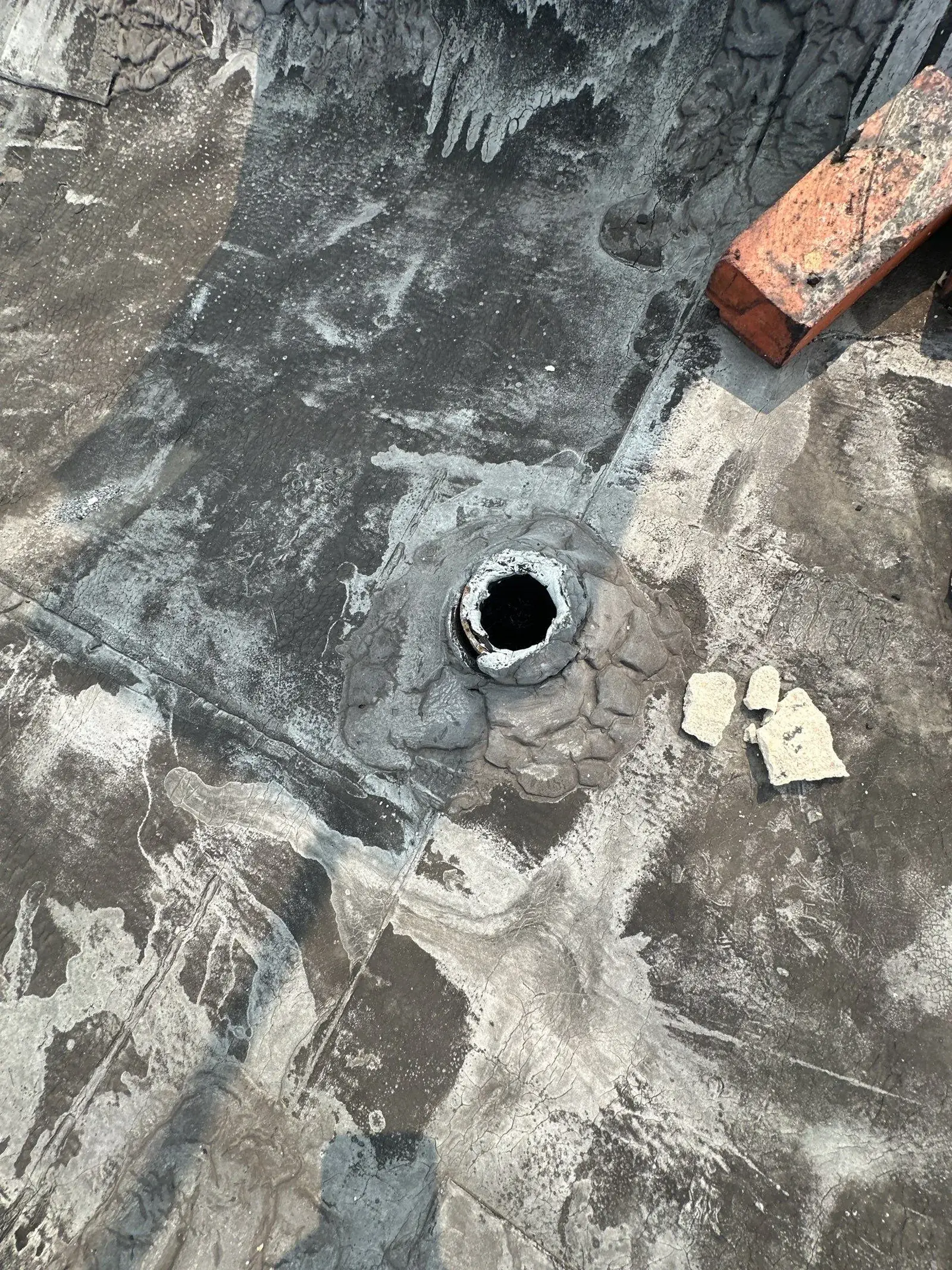 An open pipe penetration in the old coated flat roof surface, loose bricks and a cap lying beside it