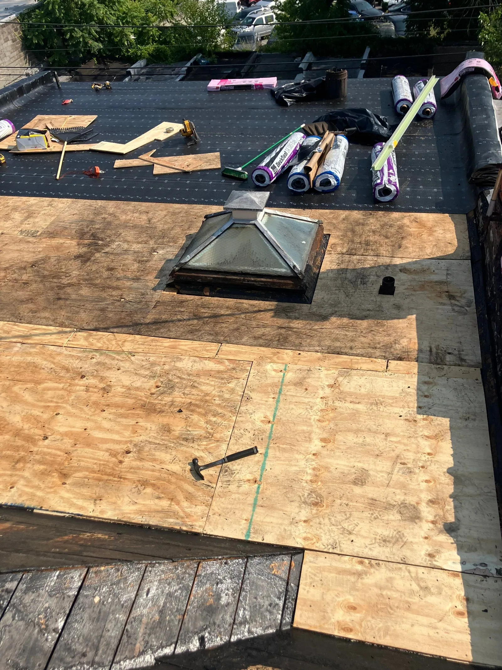New plywood sheets laid over a low-slope roof deck beside a skylight curb, with rolls of base sheet and hand tools staged on the old surface