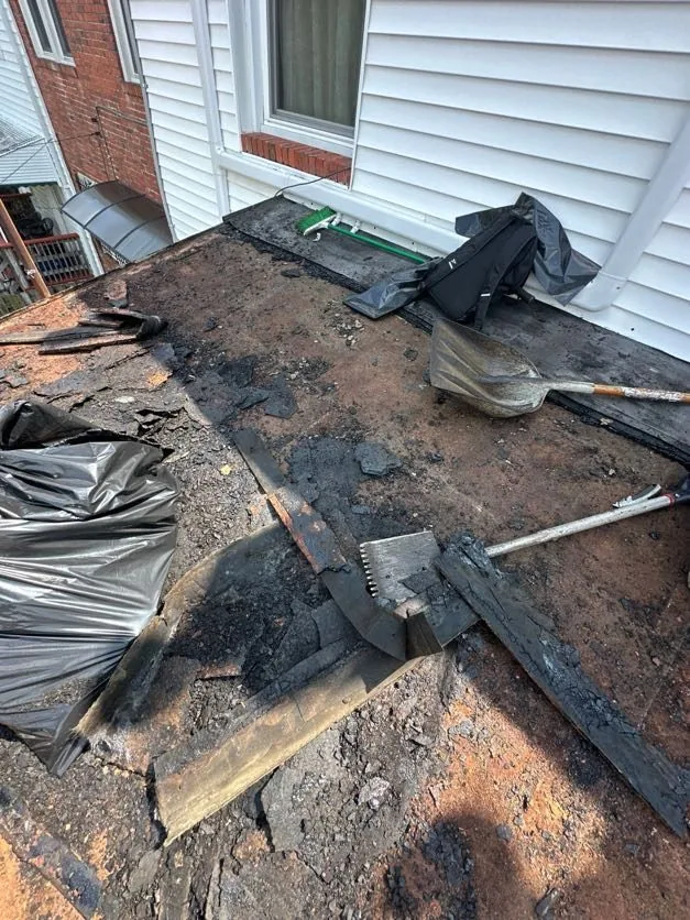 Small rear low-slope roof stripped of its old rolled roofing down to bare plank decking, with roofing spades and debris bags