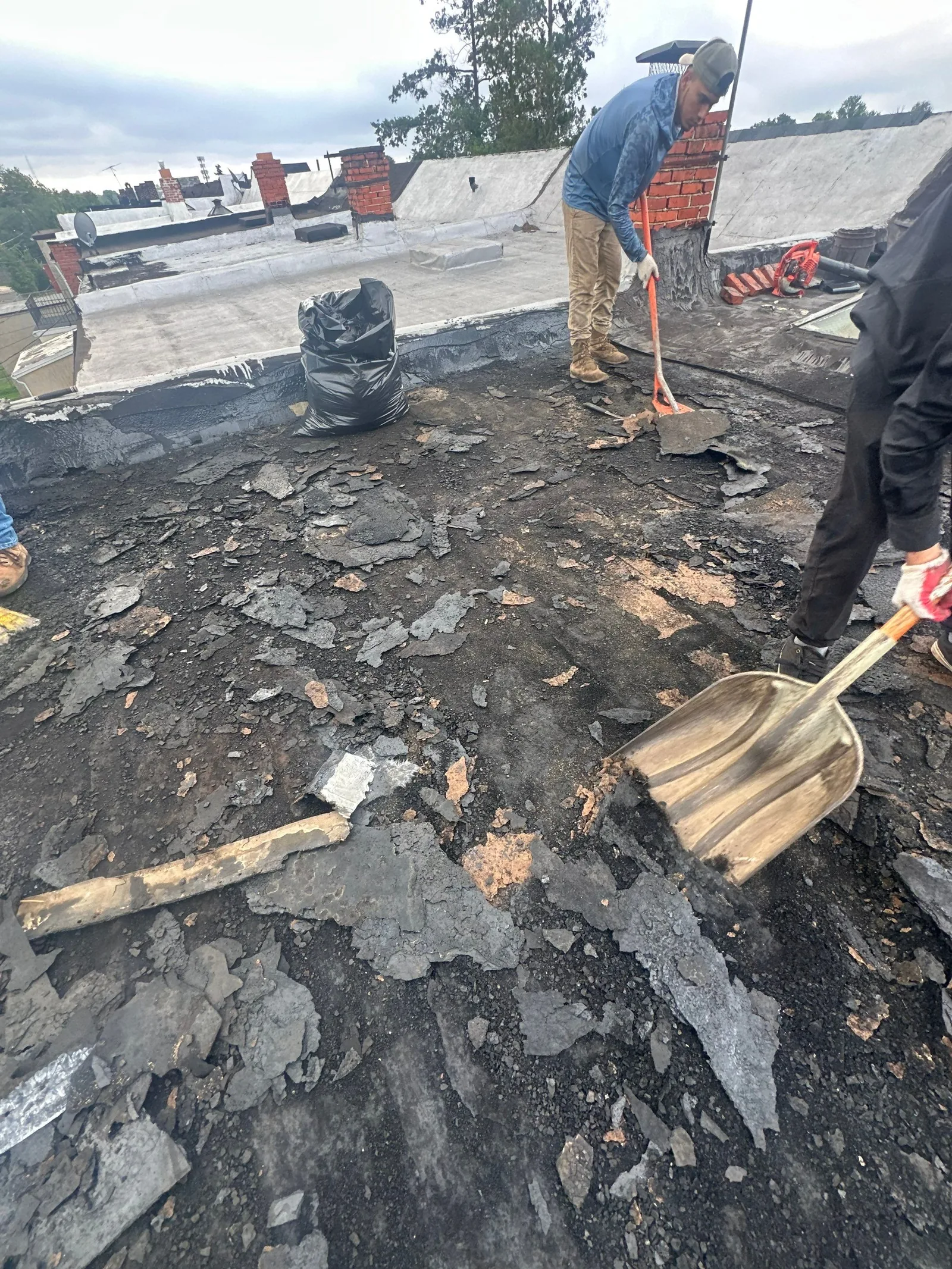 Roofer scraping old rolled roofing off a low-slope roof with a shovel, torn scraps and a debris bag on the deck, chimneys and rooftops behind