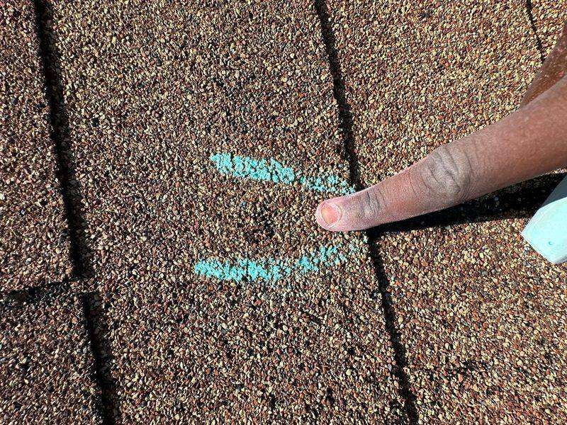 Finger pointing between two teal chalk strokes on a brown three-tab shingle, a stick of chalk at the right edge