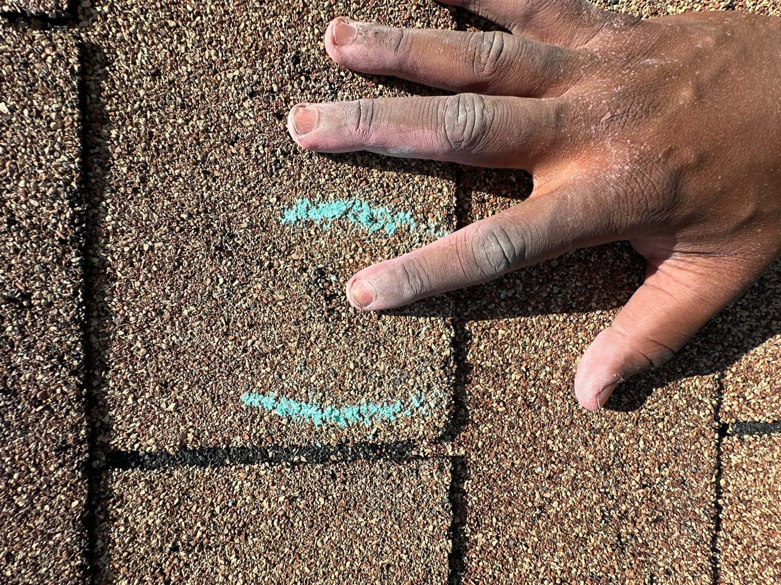 A hand laid flat on tan three-tab shingles next to two chalk-marked hail hits, for scale