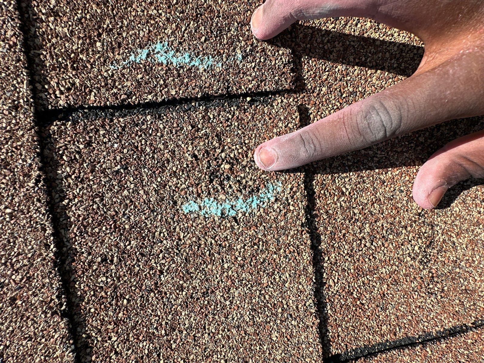 Fingers lifting the edge of a three-tab shingle next to two chalk marks