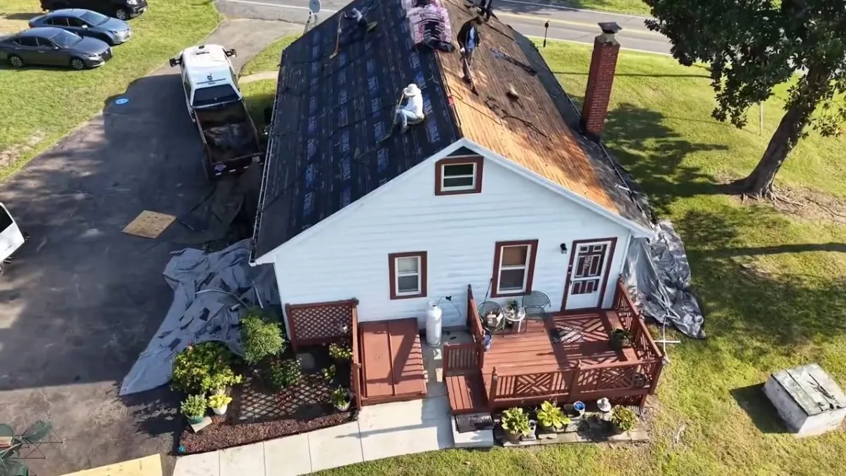 Aerial view of the rear corner of a gray roadside cottage mid-job, the near slope stripped to wood planks