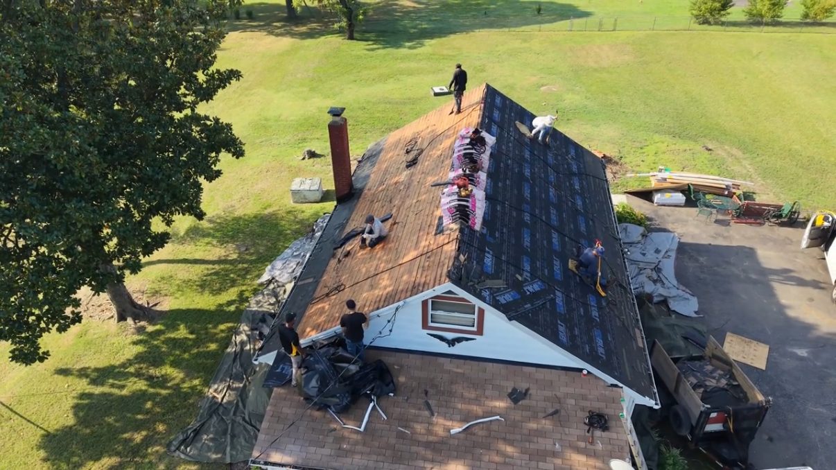 Aerial view of a house mid-replacement, one slope stripped to bare wood plank decking beside a brick chimney and the other covered in dark underlayment, a crew working and debris on the lawn