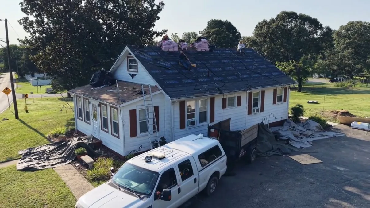 Aerial view of a gray roadside cottage mid-job, the main slope covered in dark underlayment with bundles at the ridge