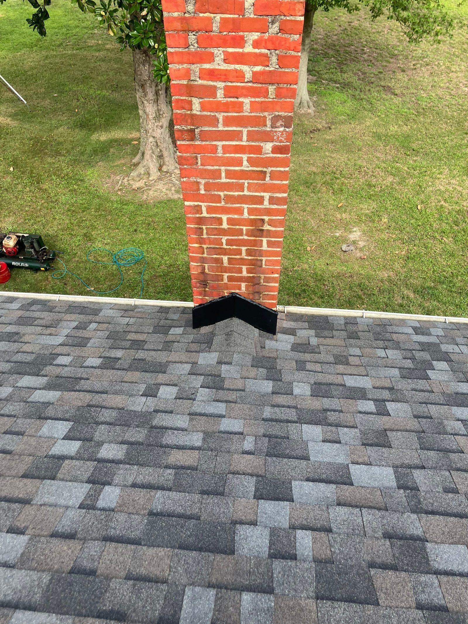 Brick chimney on a finished gray architectural shingle roof with new black base flashing, lawn and shade tree beyond