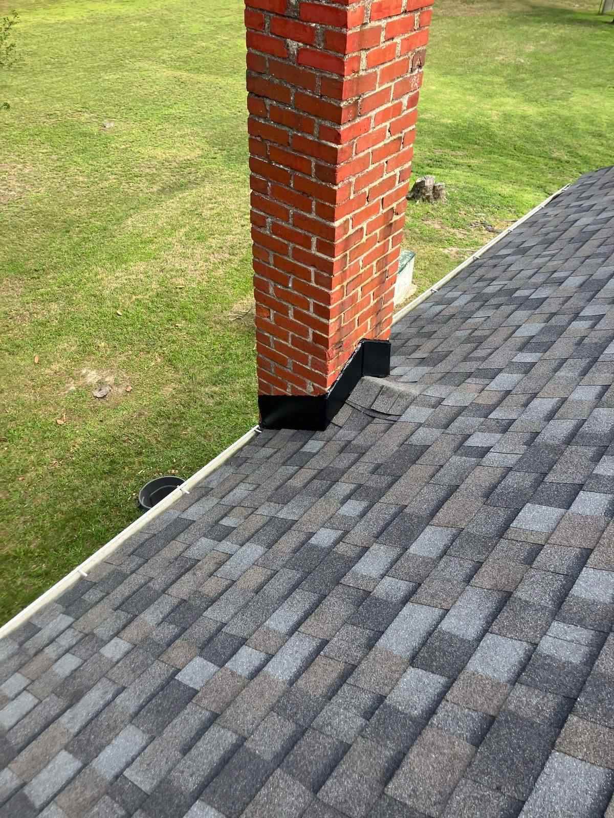 Finished gray architectural shingle slope with new black flashing tied in around a red brick chimney, green lawn beyond