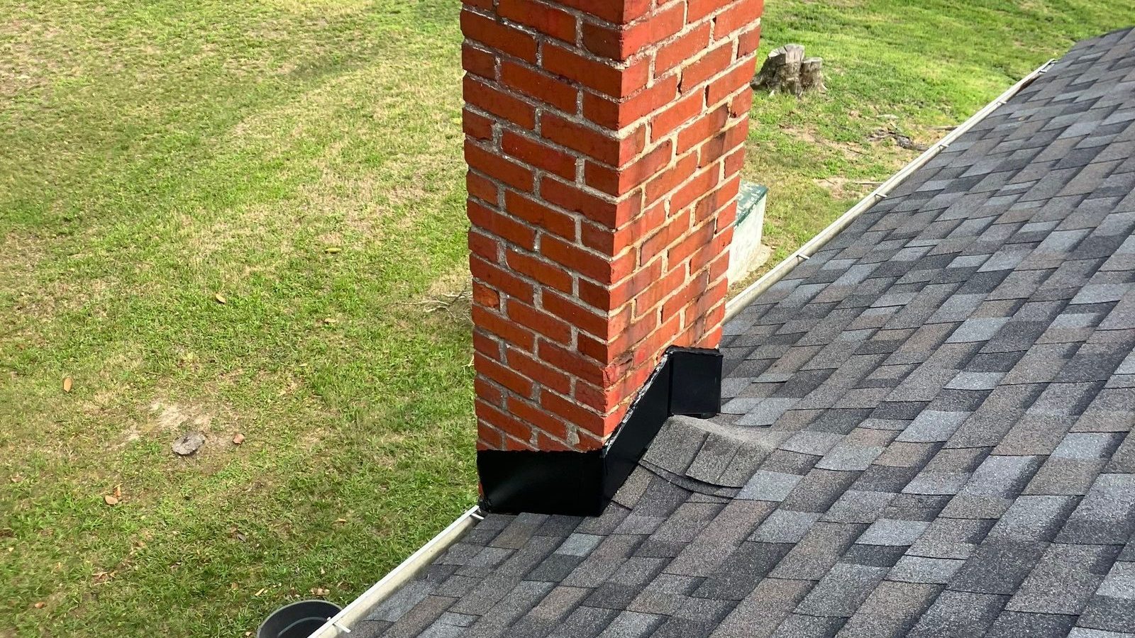 Red brick chimney rising out of a finished gray architectural shingle slope with new black base flashing