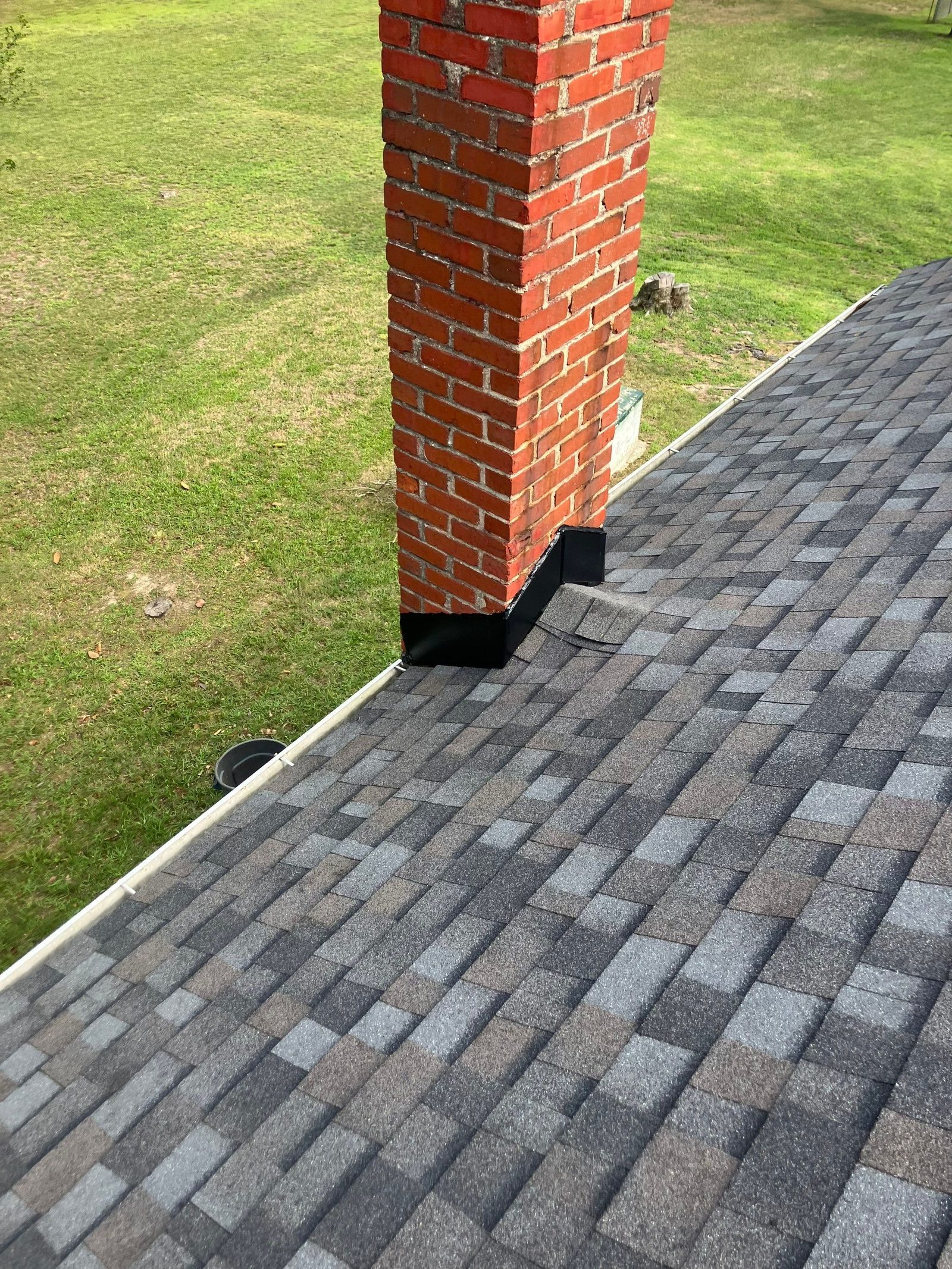 Red brick chimney rising out of a finished gray architectural shingle slope with new black base flashing