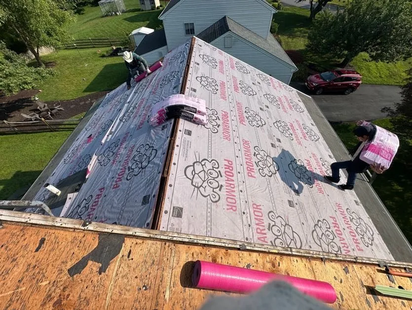 Steep hip roof fully covered in printed synthetic underlayment, with two roofers carrying shingle bundles up the slope