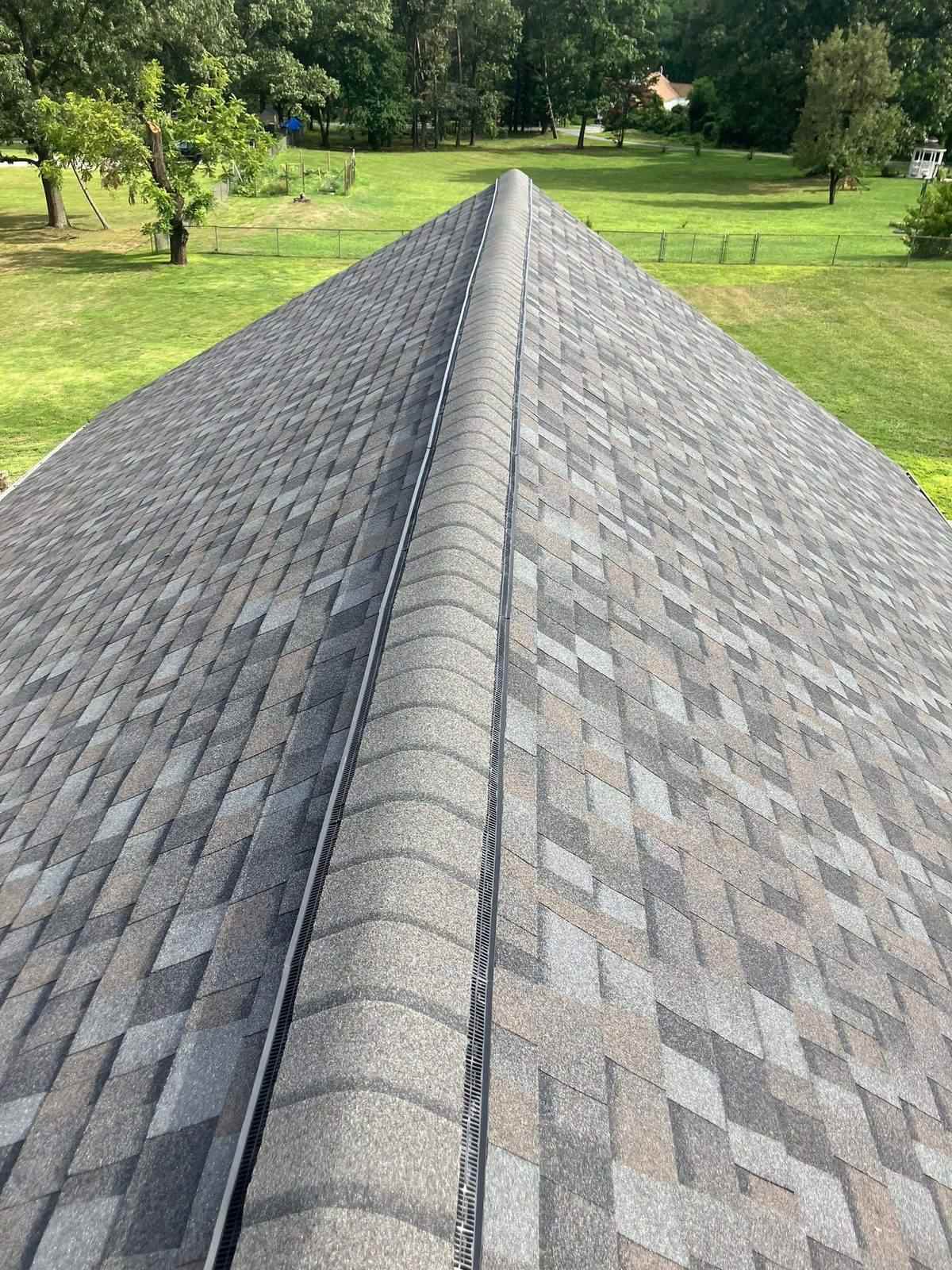 Two gray shingle slopes meeting at the ridge with a continuous ridge vent installed along the peak, back lawn and trees behind