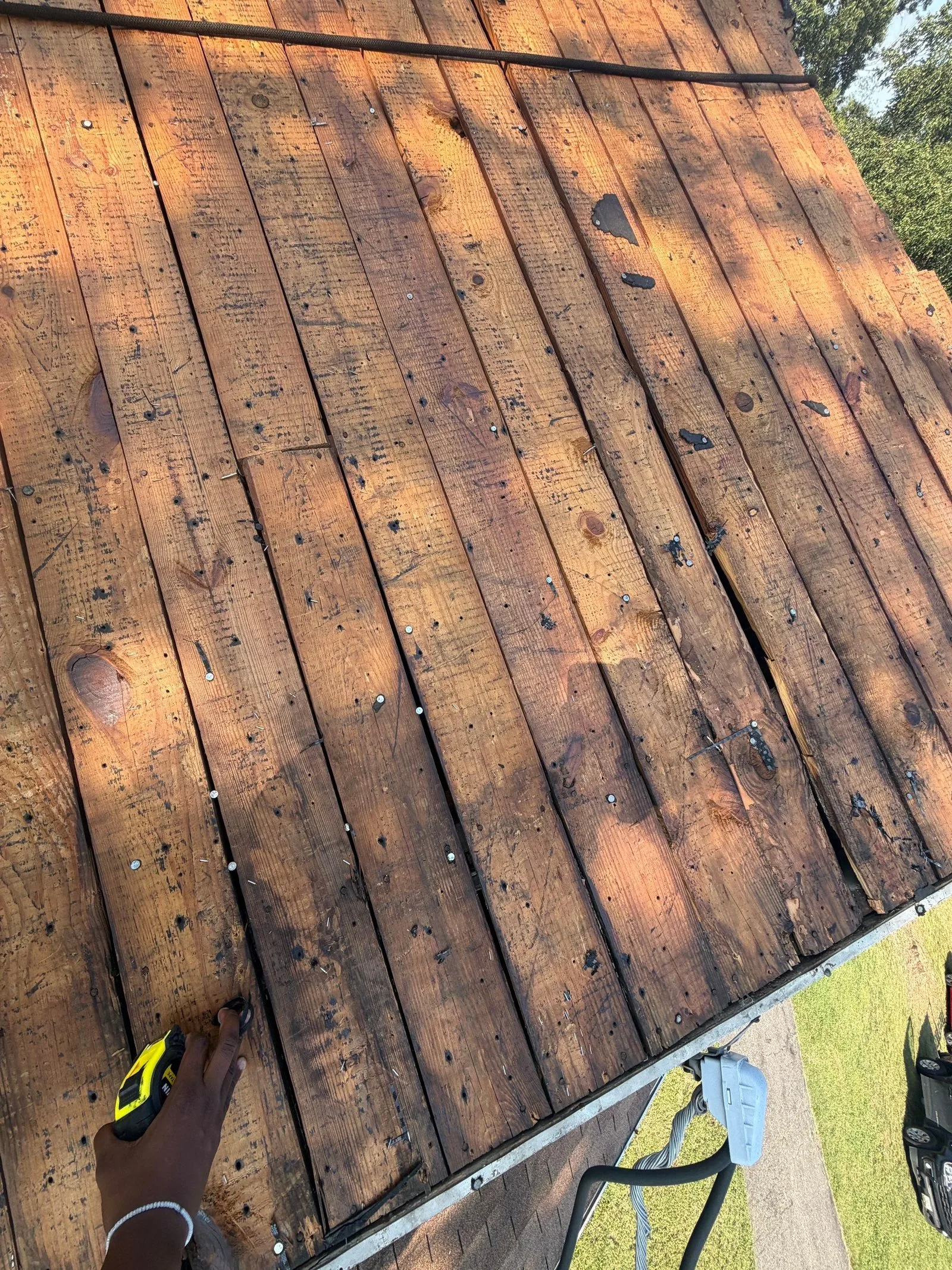 Wide sweep of bare plank decking studded with old nails, a hand holding a tape measure at the lower left