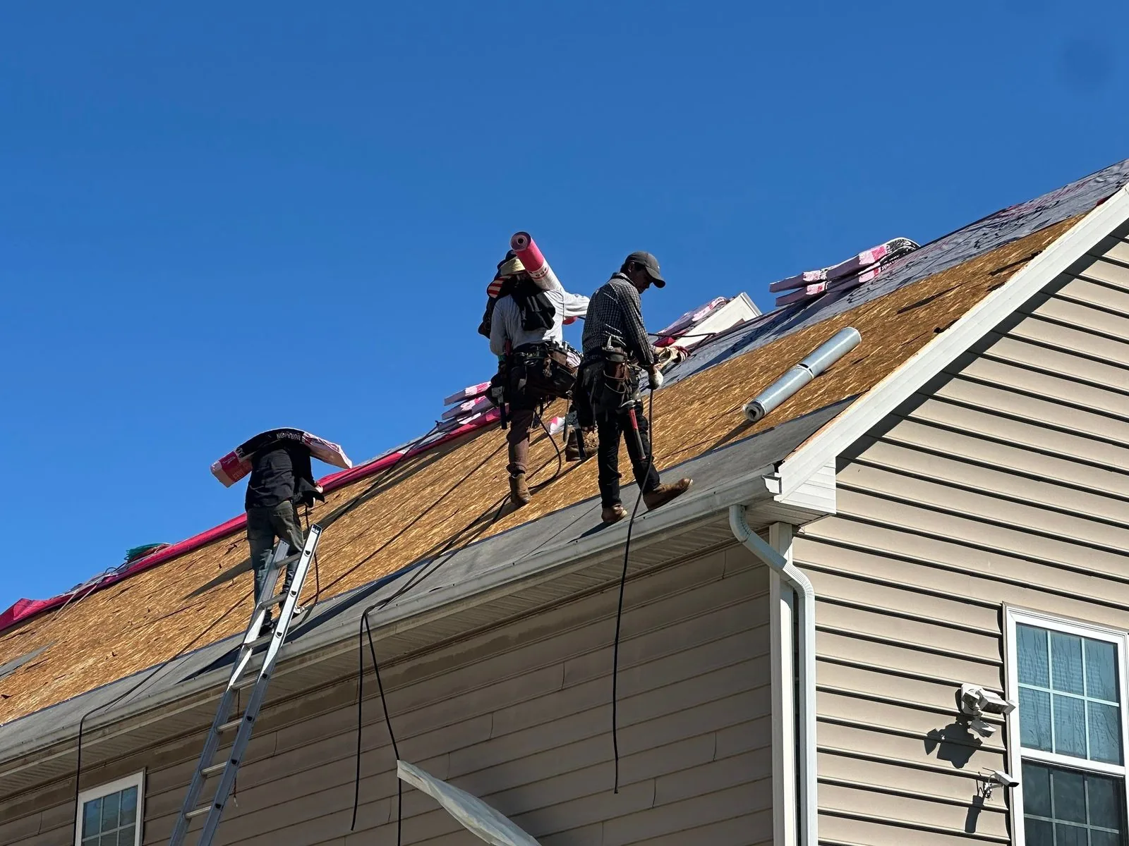 Three roofers on the stripped OSB deck of a large tan vinyl-sided house against a deep blue sky