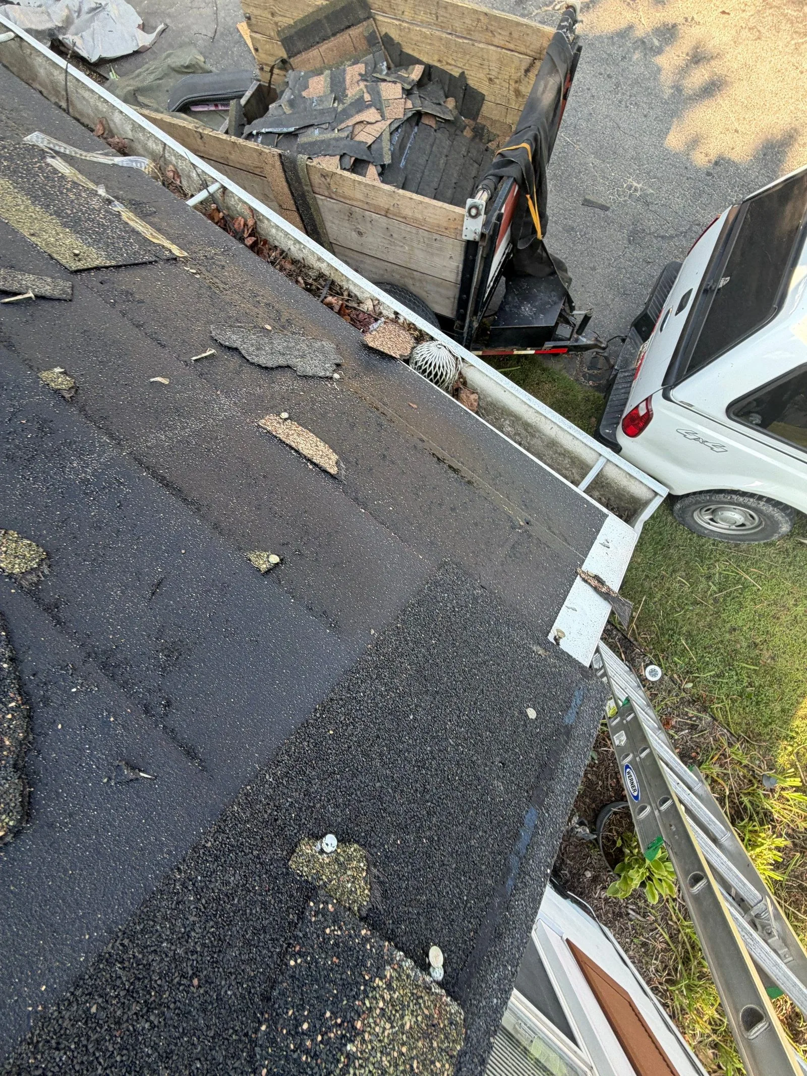 Looking down the eave at tear-off debris caught in the gutter, with a dump trailer heaped with old shingles below