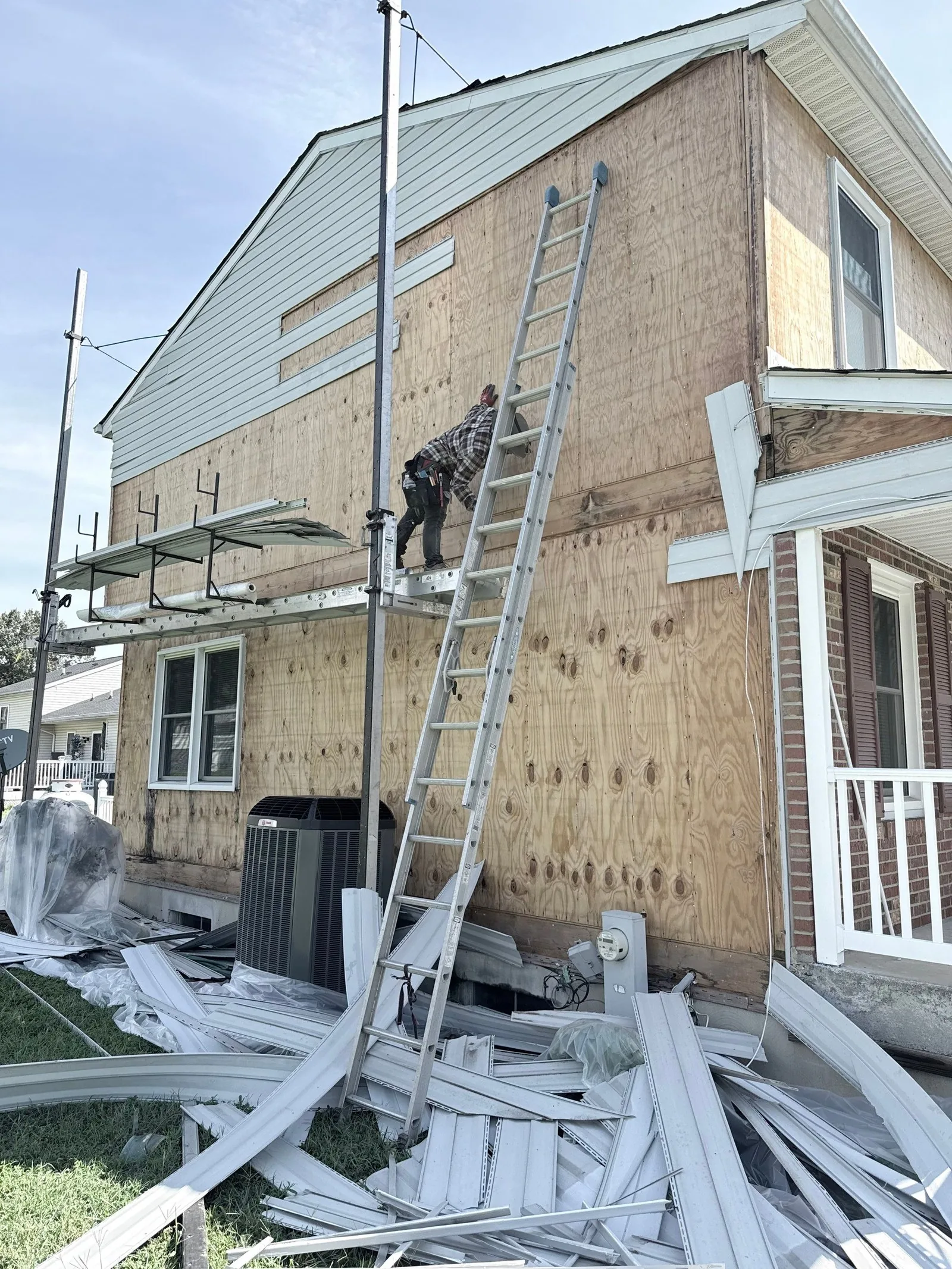 Two-storey wall stripped to plywood while a roofer works from pump-jack staging above the removed siding