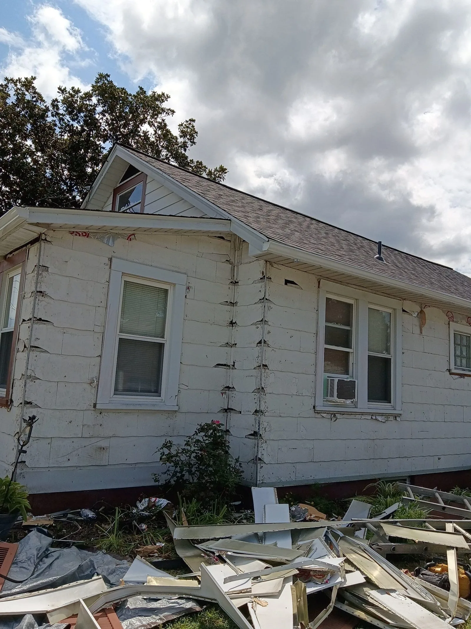 Side of the house with the white siding partly stripped off, torn siding and trim piled on the ground below