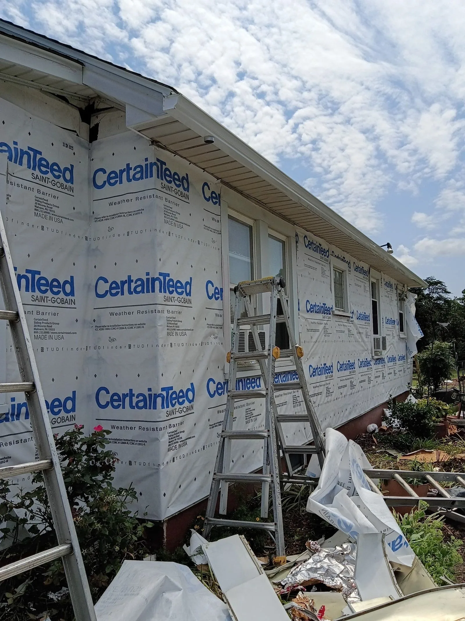 Side of the house stripped back to the house wrap, ladders leaning against it and torn siding scattered on the ground